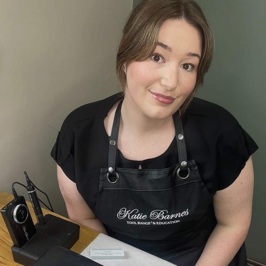 Woman wearing a black apron with 'Katie Barnes' on it, sitting at a desk.
