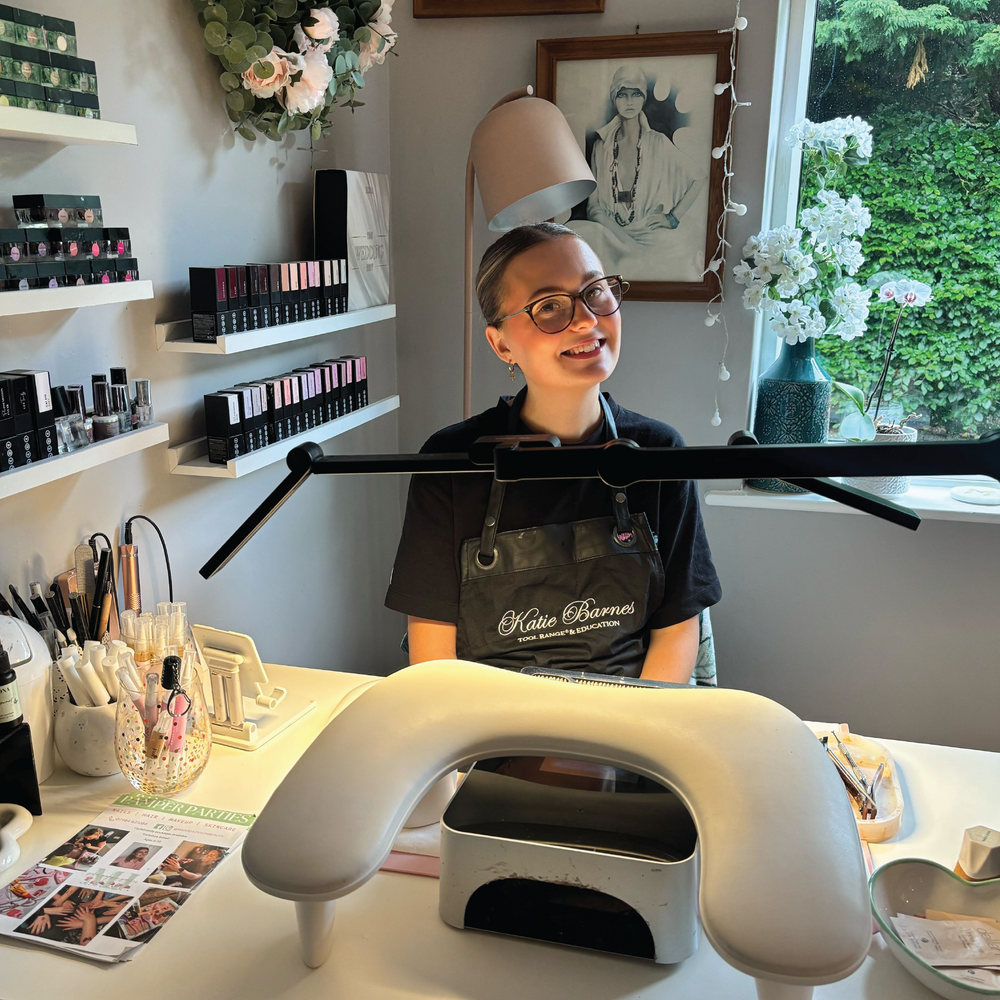 Nail Technician sitting at a nail salon desk with KB Flexible Desk Light and wearing Katie Barnes Tool Range apron.