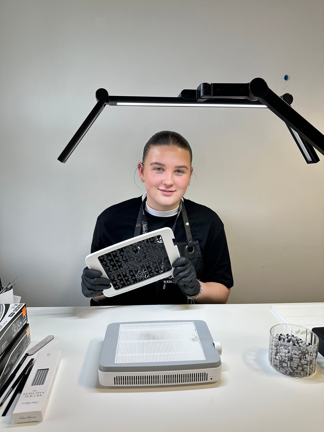 woman with brown hair in a bun, holding the grill of Katie Barnes dust collector. Katie Barnes black clamp desk lamp can be seen over the nail technicians head.